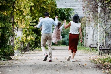 Familie mit einem Kind, das zwischen Eltern in einer grünen Allee schwingt.